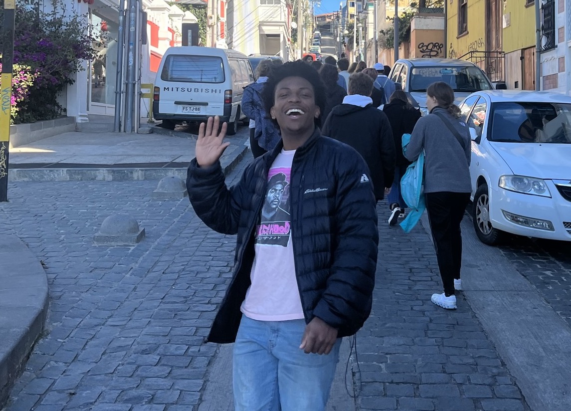 A young man waves and smiles at the camera while standing on a street in Santiago, Chile