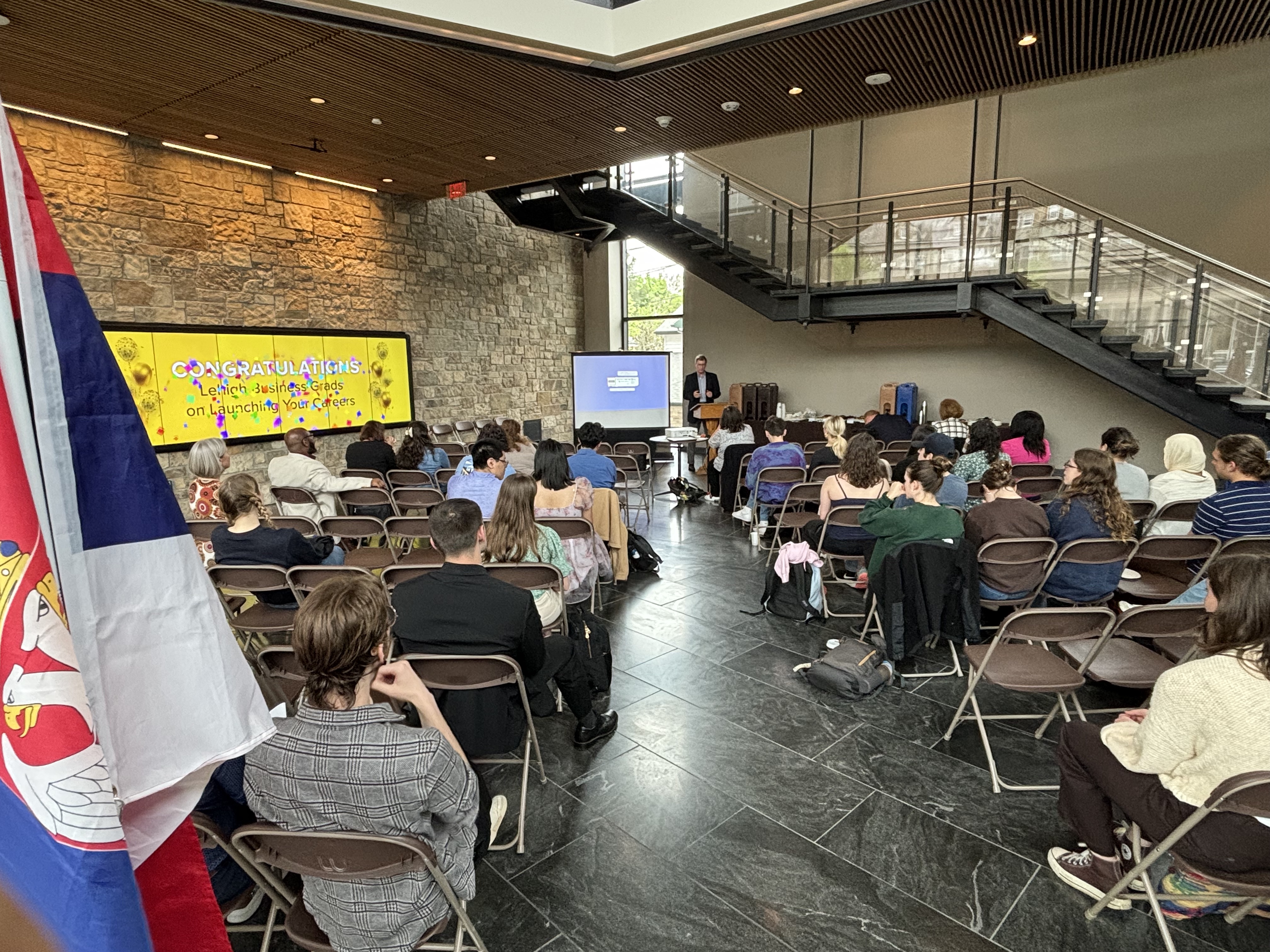 Lehigh University students in fold-out chairs inside the lobby of a building, listening to someone speak at a podium