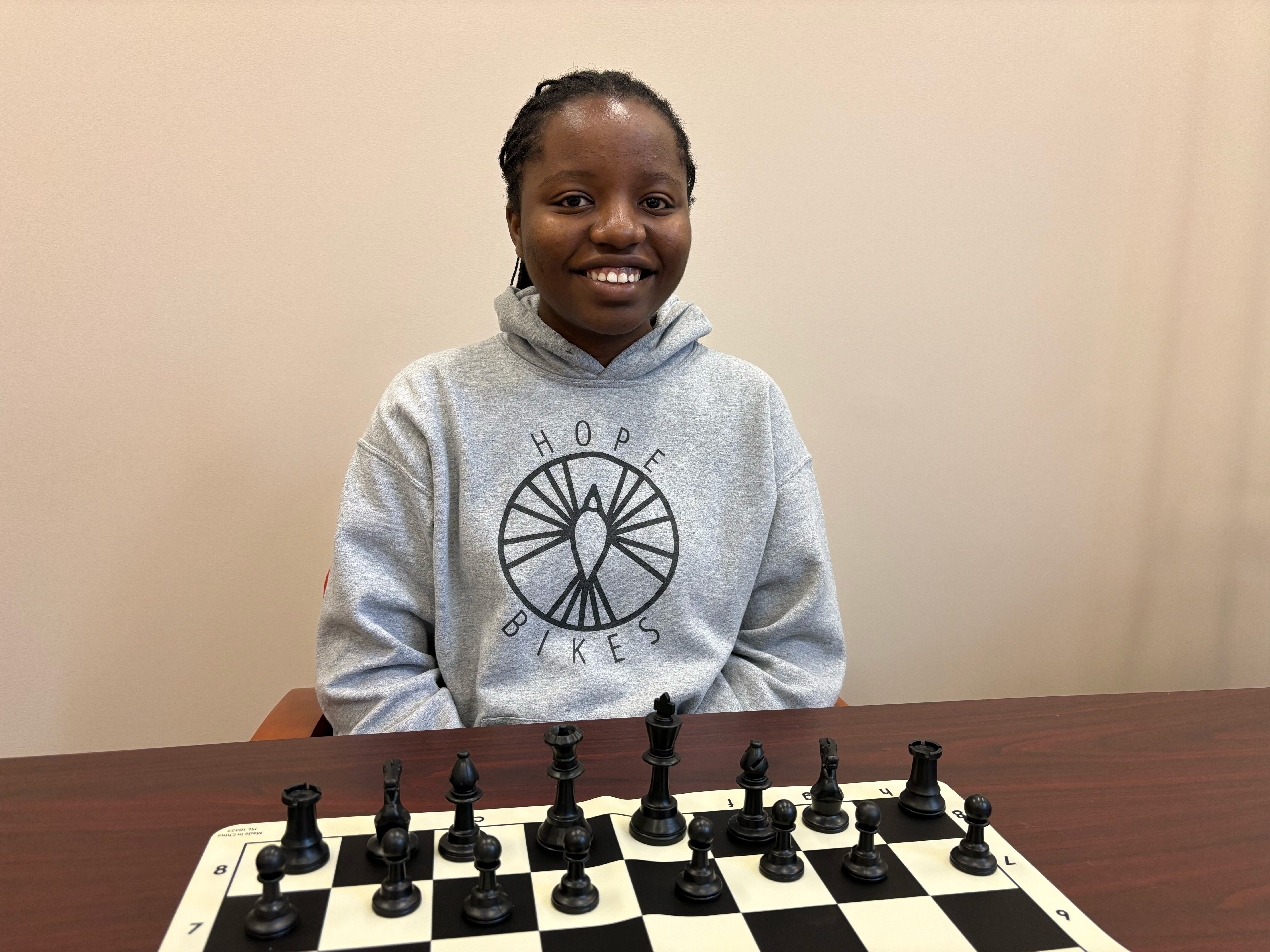 Besa Masaiti smiling for the camera, sitting in front of a chessboard