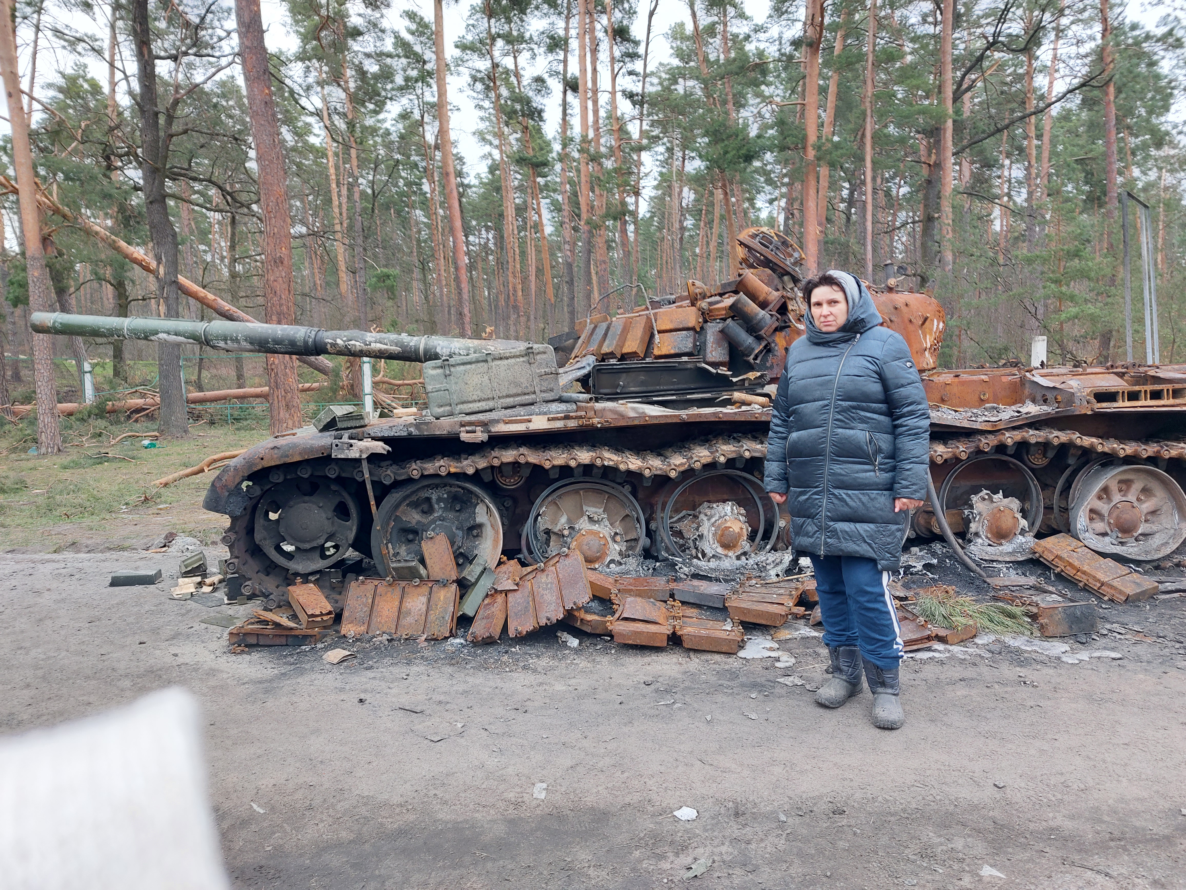 Roman Moskalenkos mother standing in front of a destroyed tank on her street in Ukraine