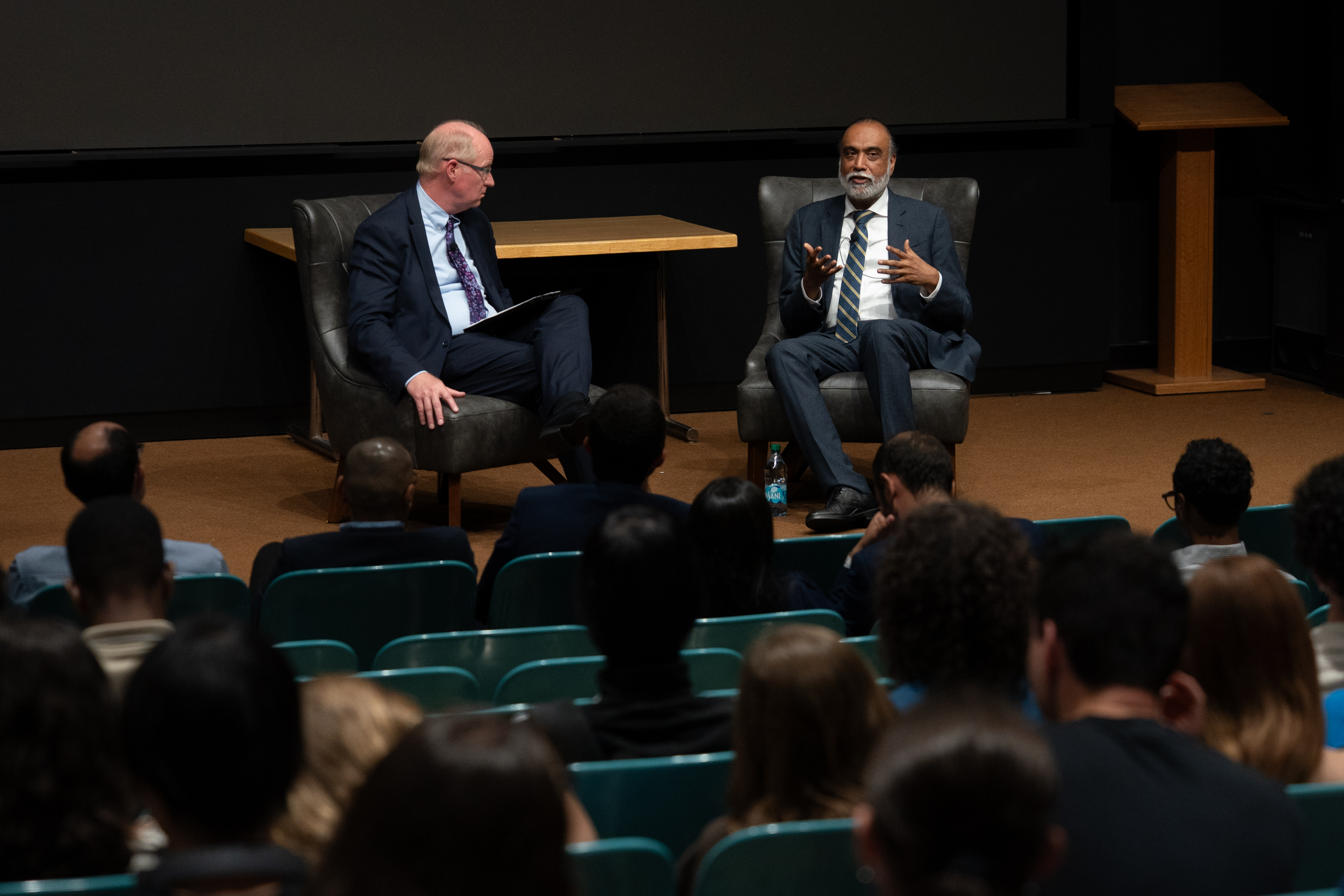 Two men sitting in chairs and speaking on a stage to an auditorium of students