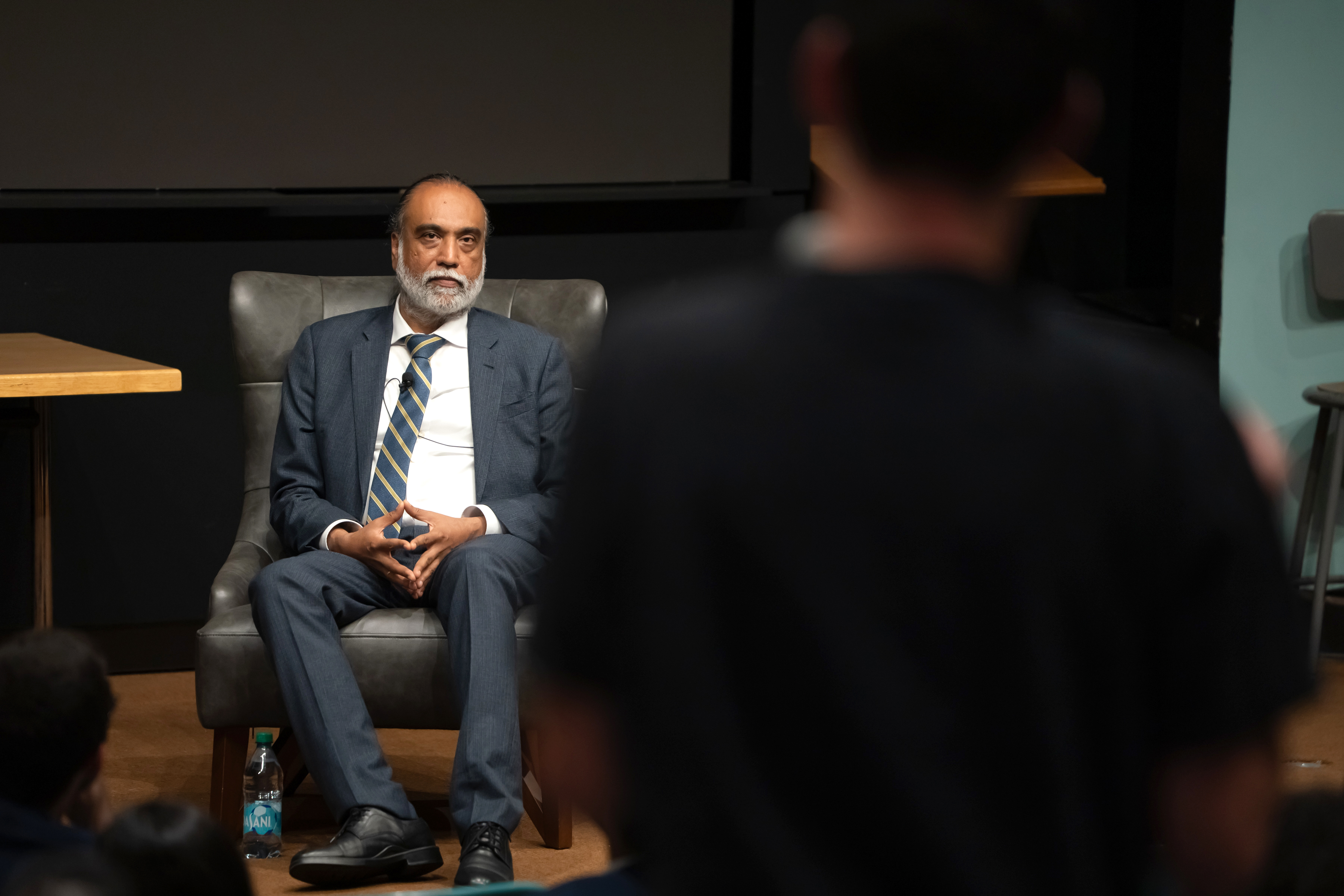 A man sitting on a chair on a stage listening to a person speaking from the audience