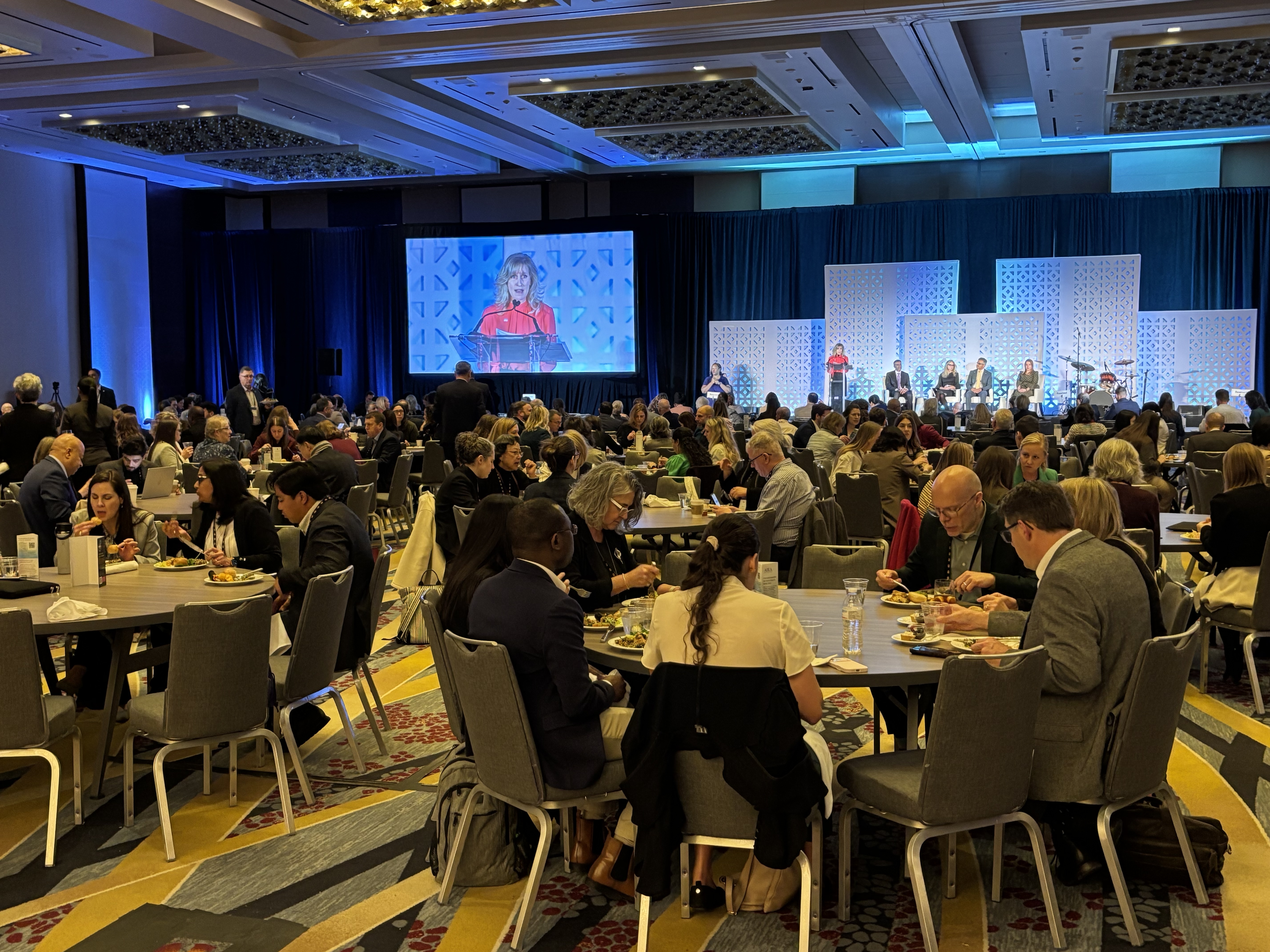 A large group of people eating at multiple tables while listening to spekaers on a stage
