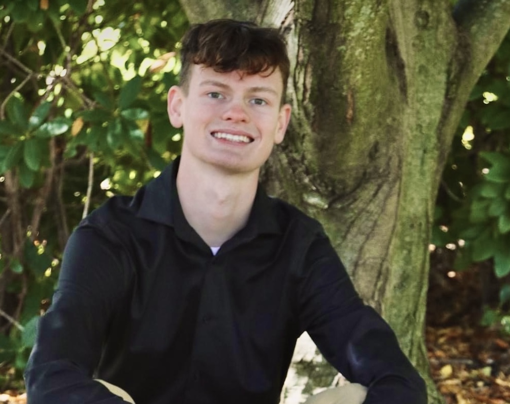 A young man smiling while sitting in front of a tree