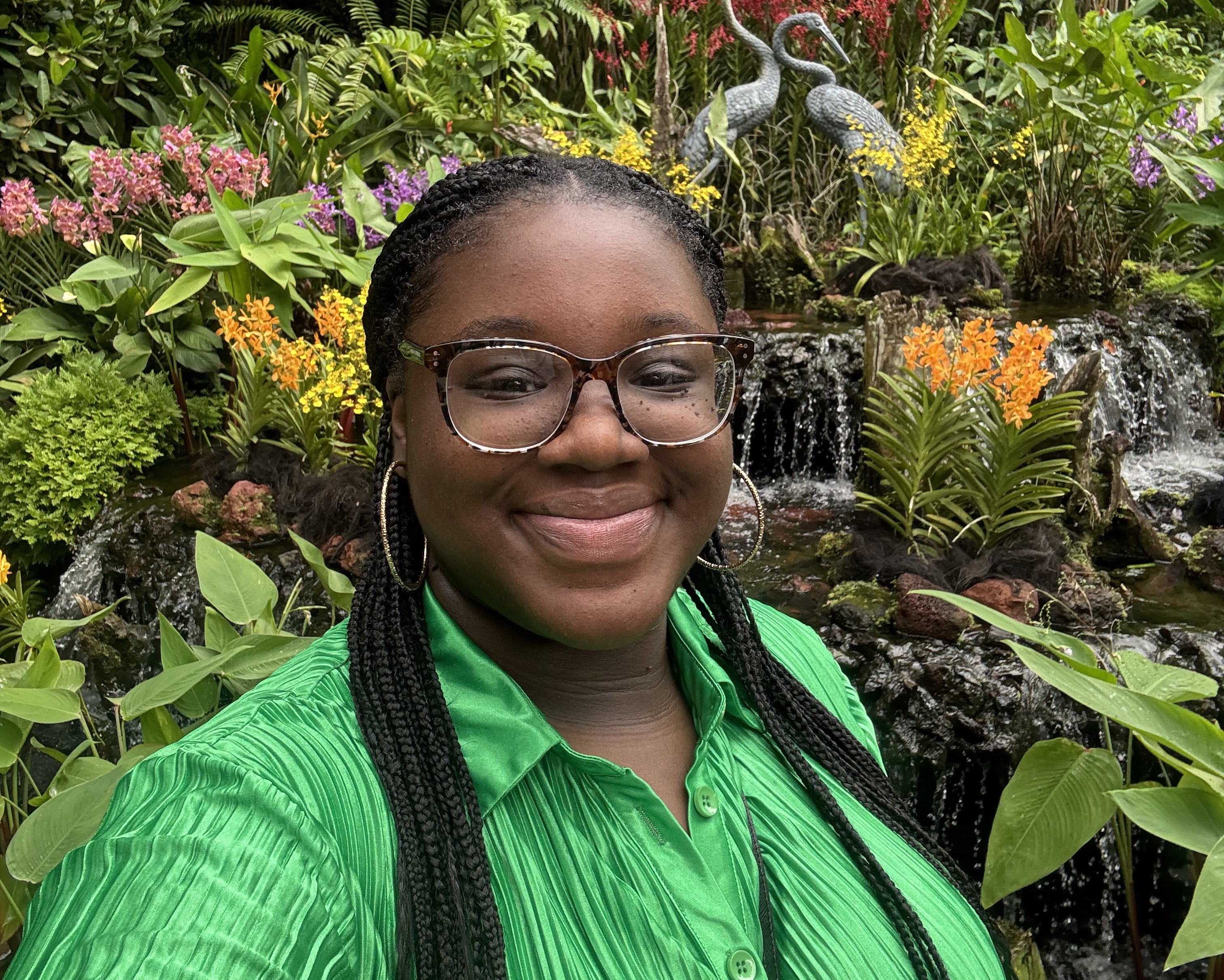 A young woman smiling for the camera with a fountain and various plants behind her