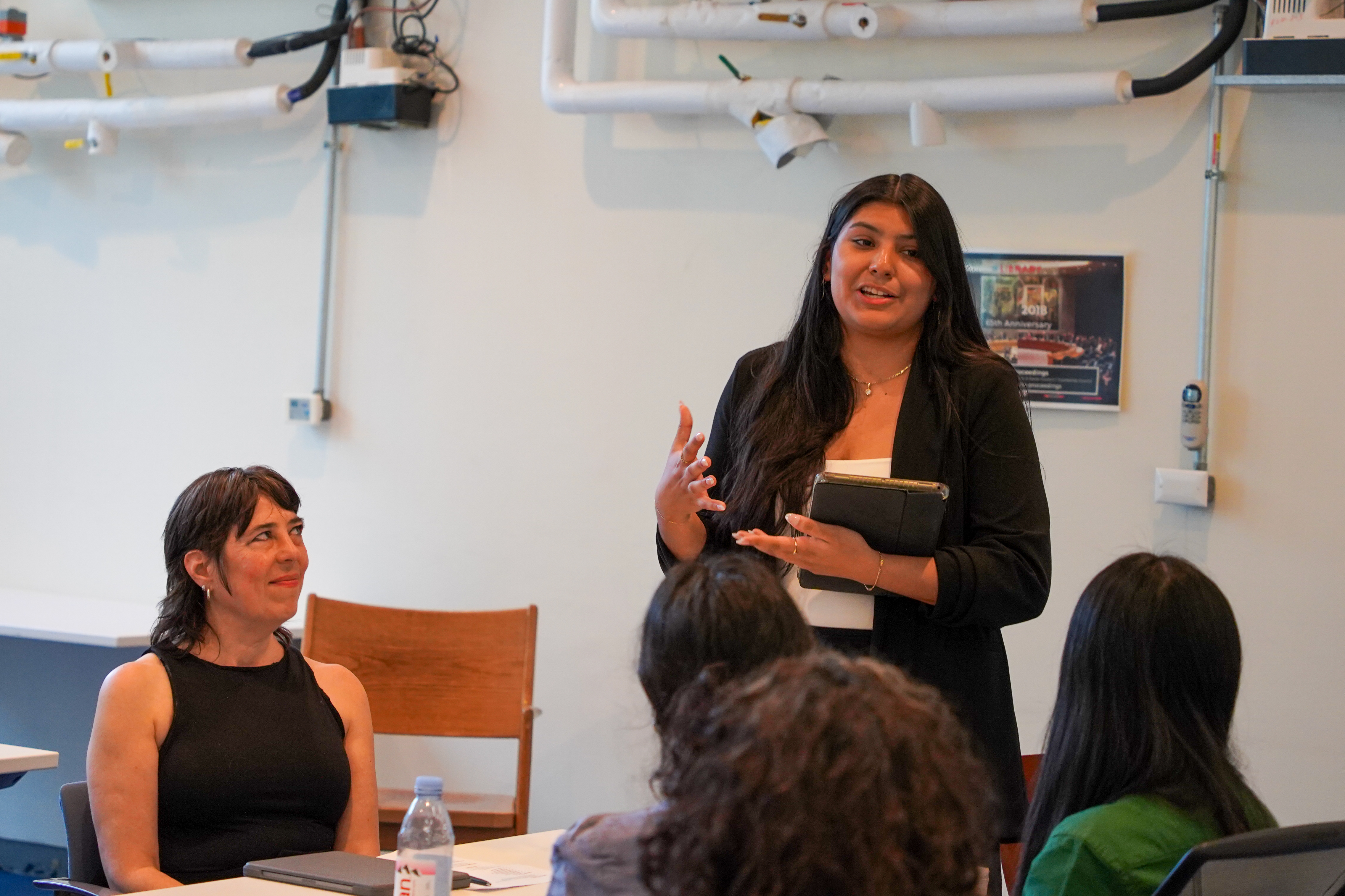 A young woman standing at a table and speaking to others in the audience