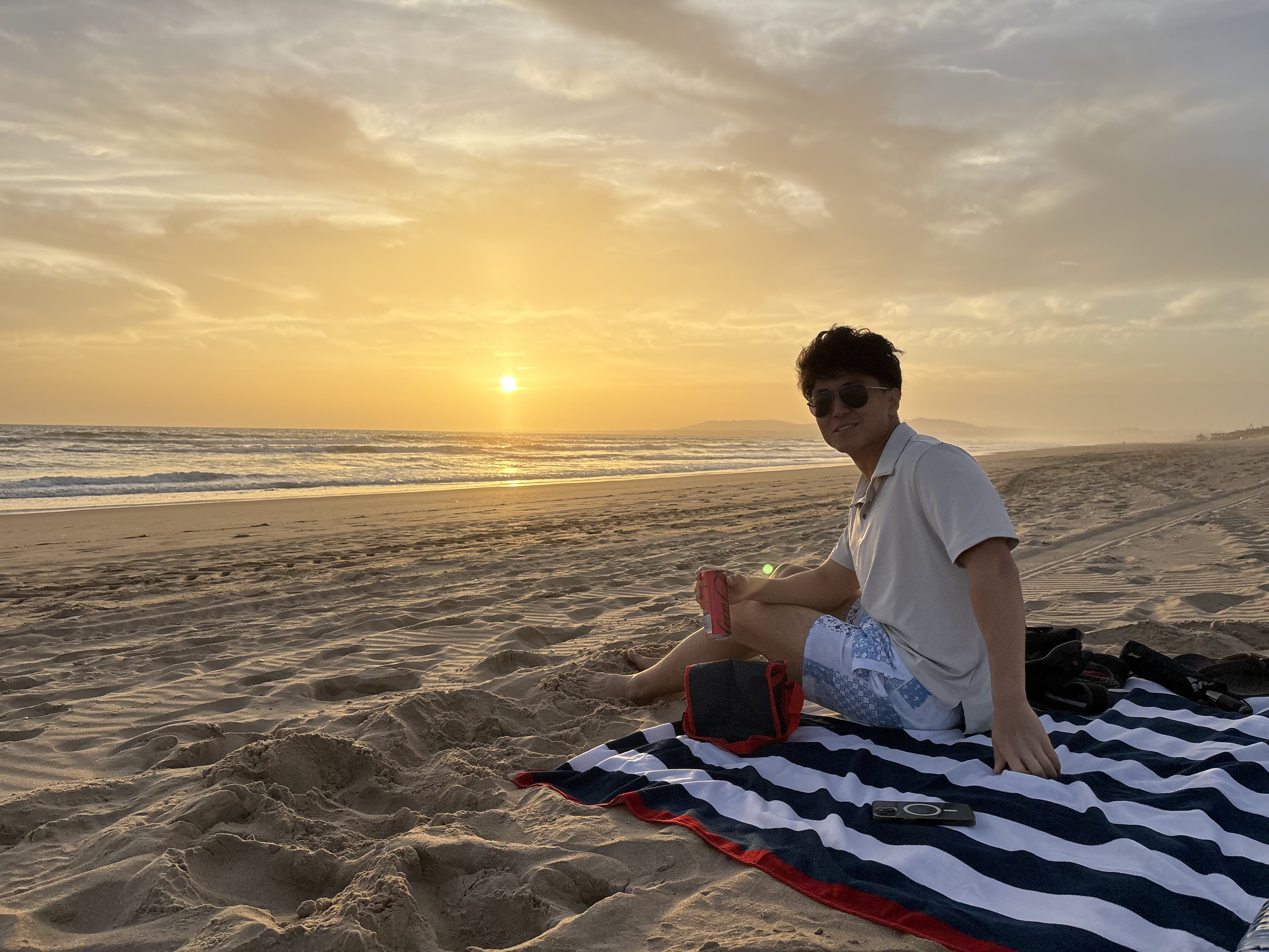 A young man sitting on a towel on a beach with a sunset behind him
