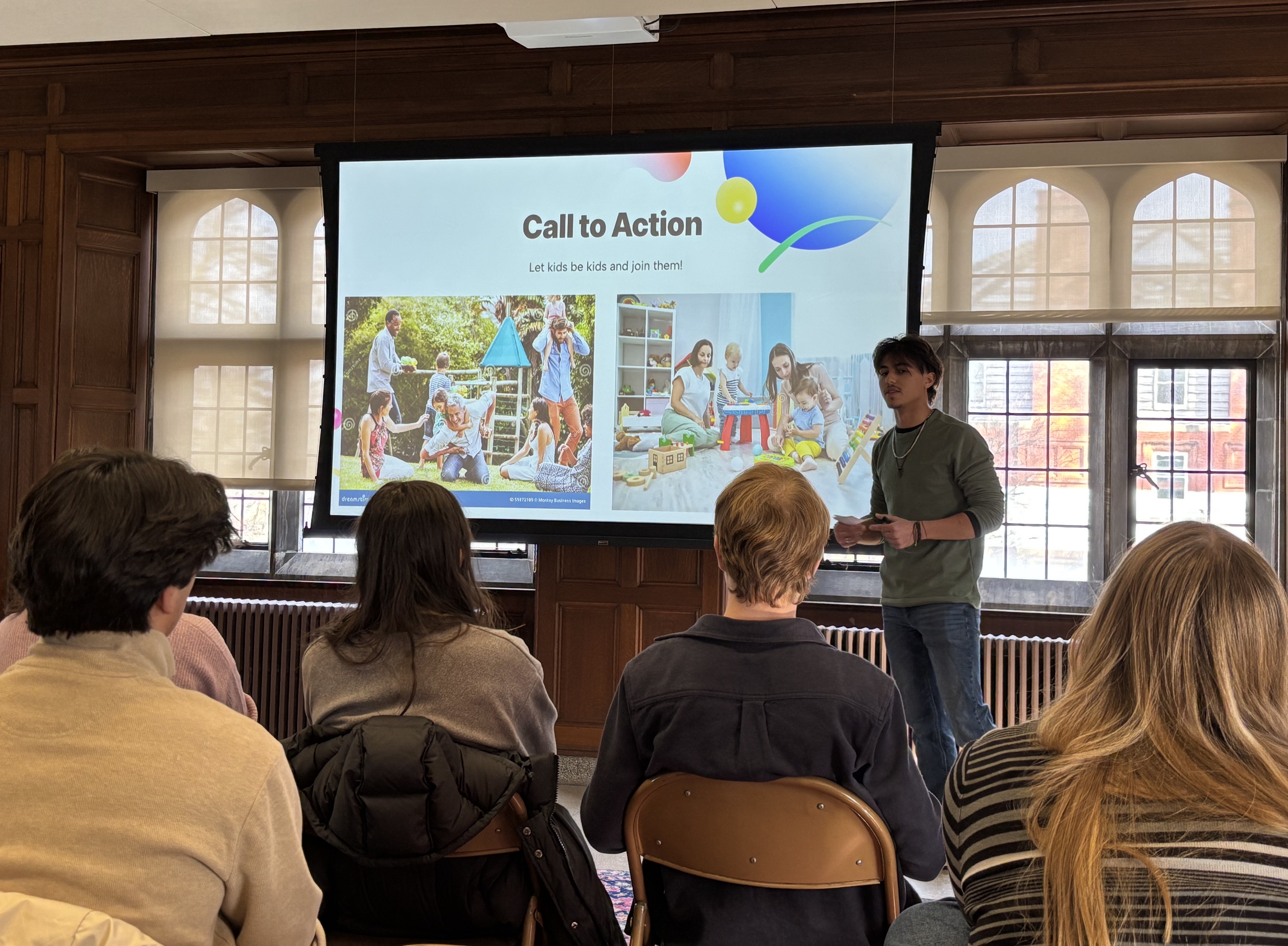 A young man standing in front of a screen, giving a presentation to a seated audience