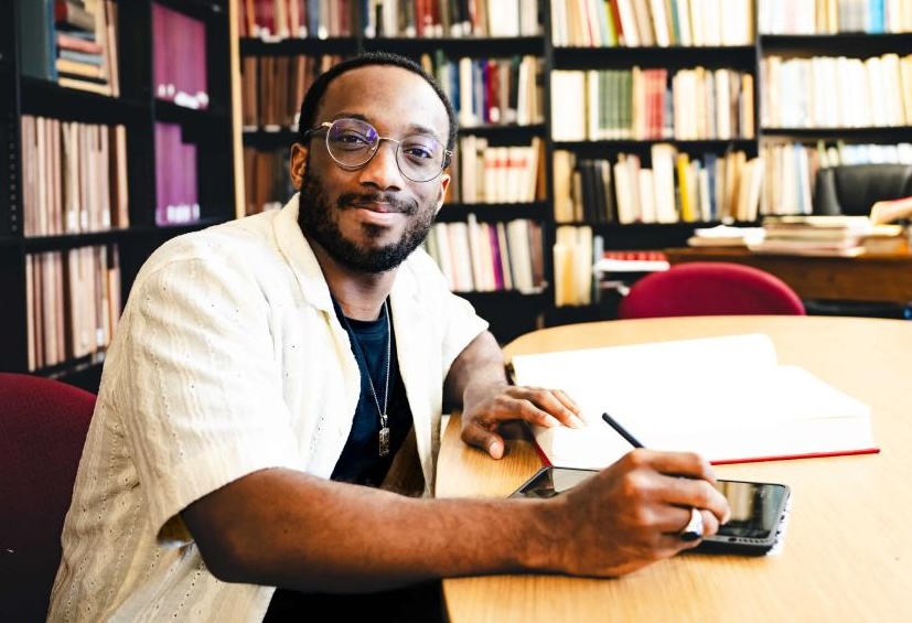 A young man sitting at a table, holding a pen and tablet with a book open in front of him