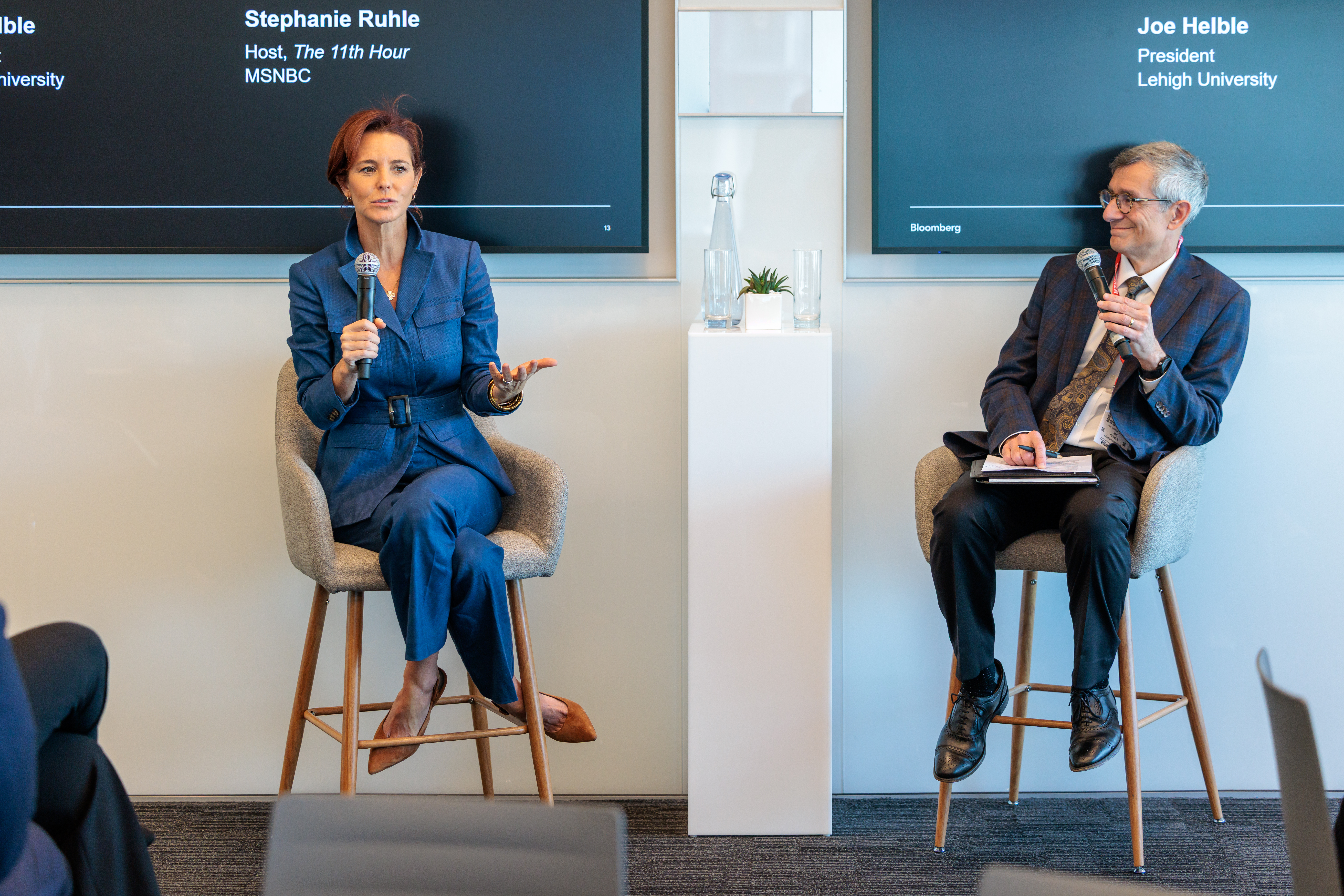 A woman and man sit on stools while talking to a crowd.