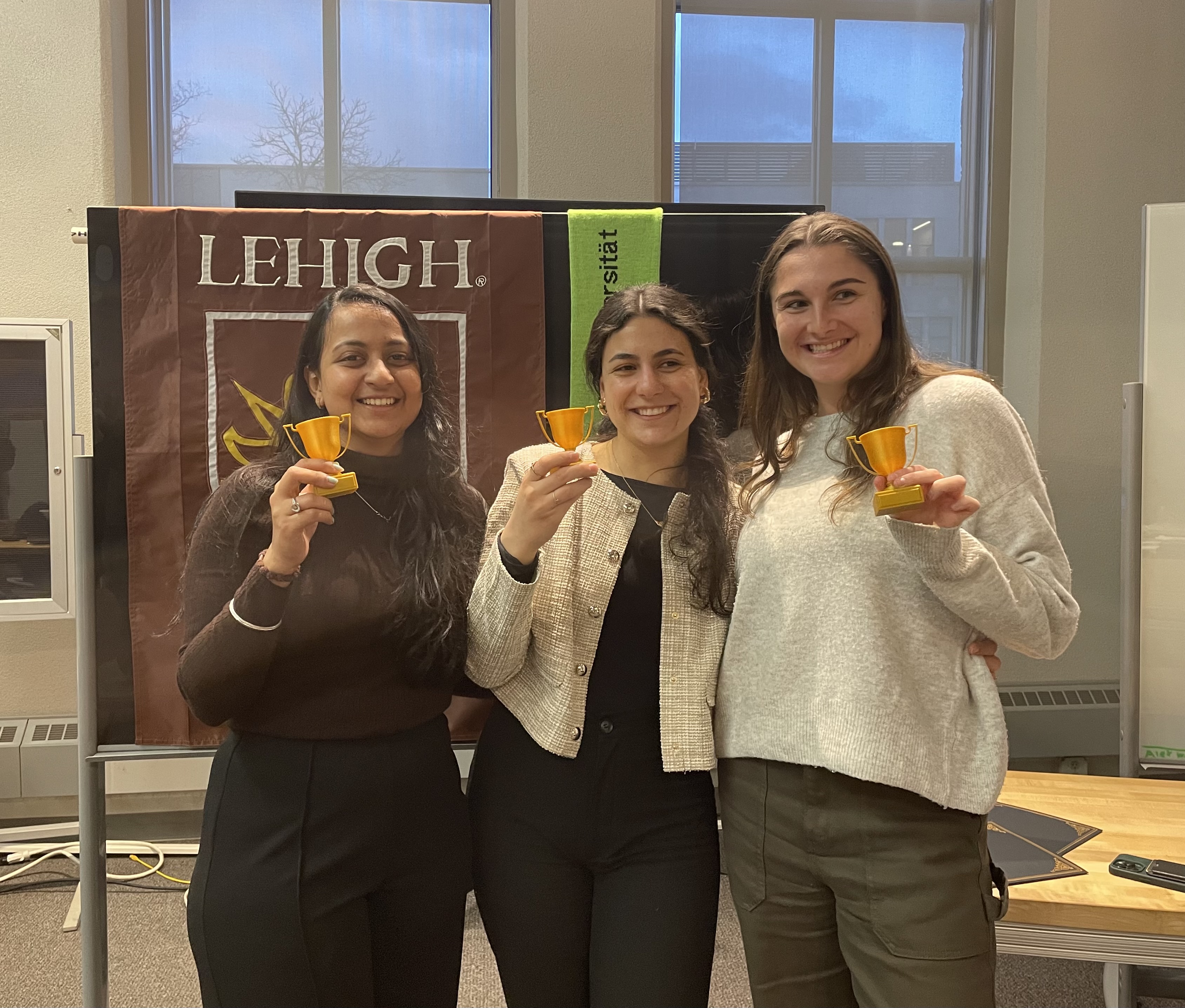 Three young women holding very small trophies in front of a Lehigh University banner