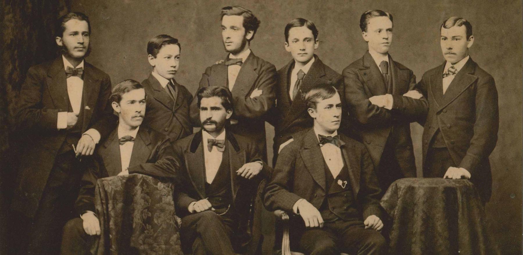A sepia-tinted photo of nine men posing for a photo in suits