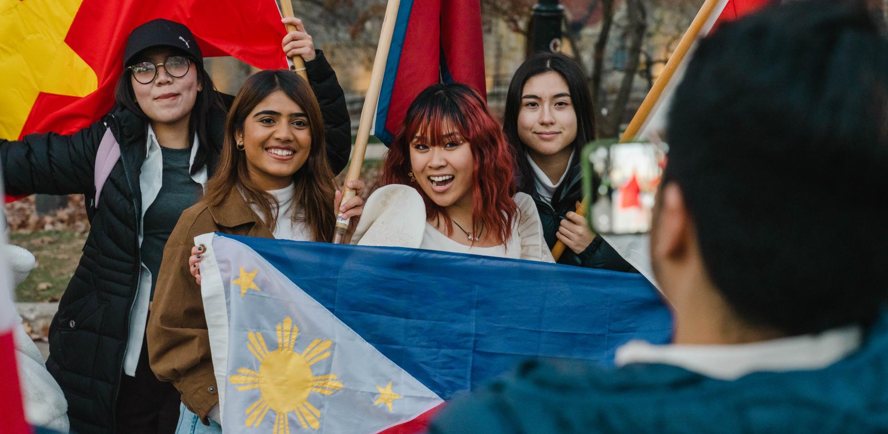 A man with his back to the camera takes a smartphone photo of four young owmen smiling and holding up a variety of flags