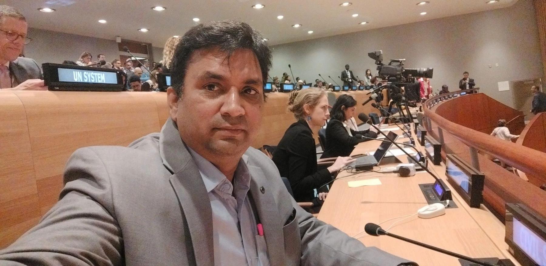 Binod Anand sitting at a microphone in a crowded room in the United Nations headquarters