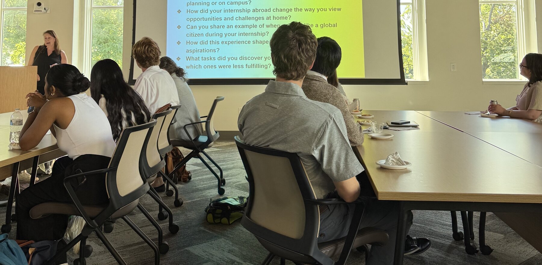 A group of people sitting at tables listening to a woman give a Powerpoint presentation