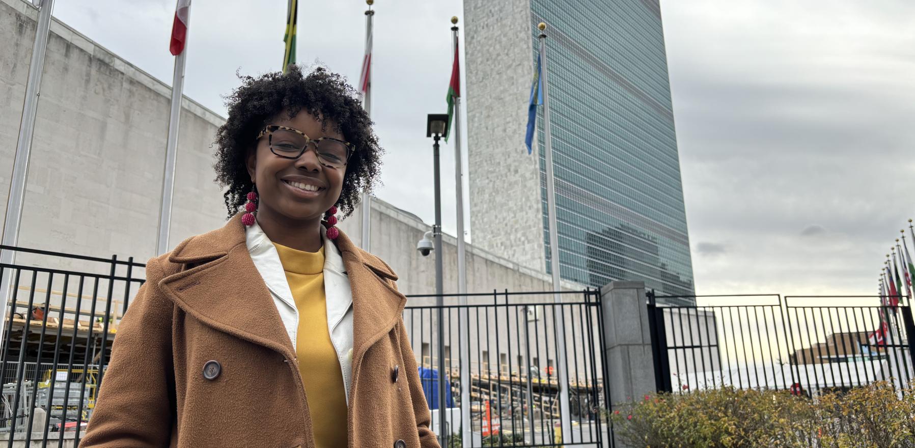 Rhema Hooper standing in front of the United Nations headquarters, wearing a tan coat, smiling