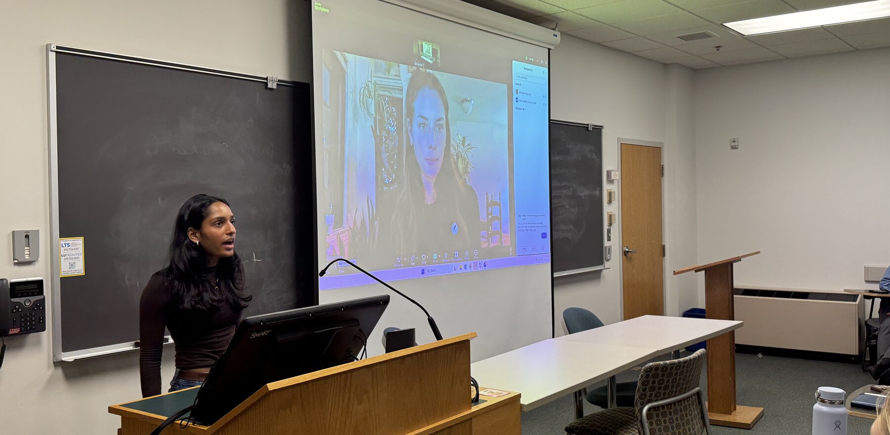 A young woman speaks at a podium while another woman speaks on an overhead projector screen behind her