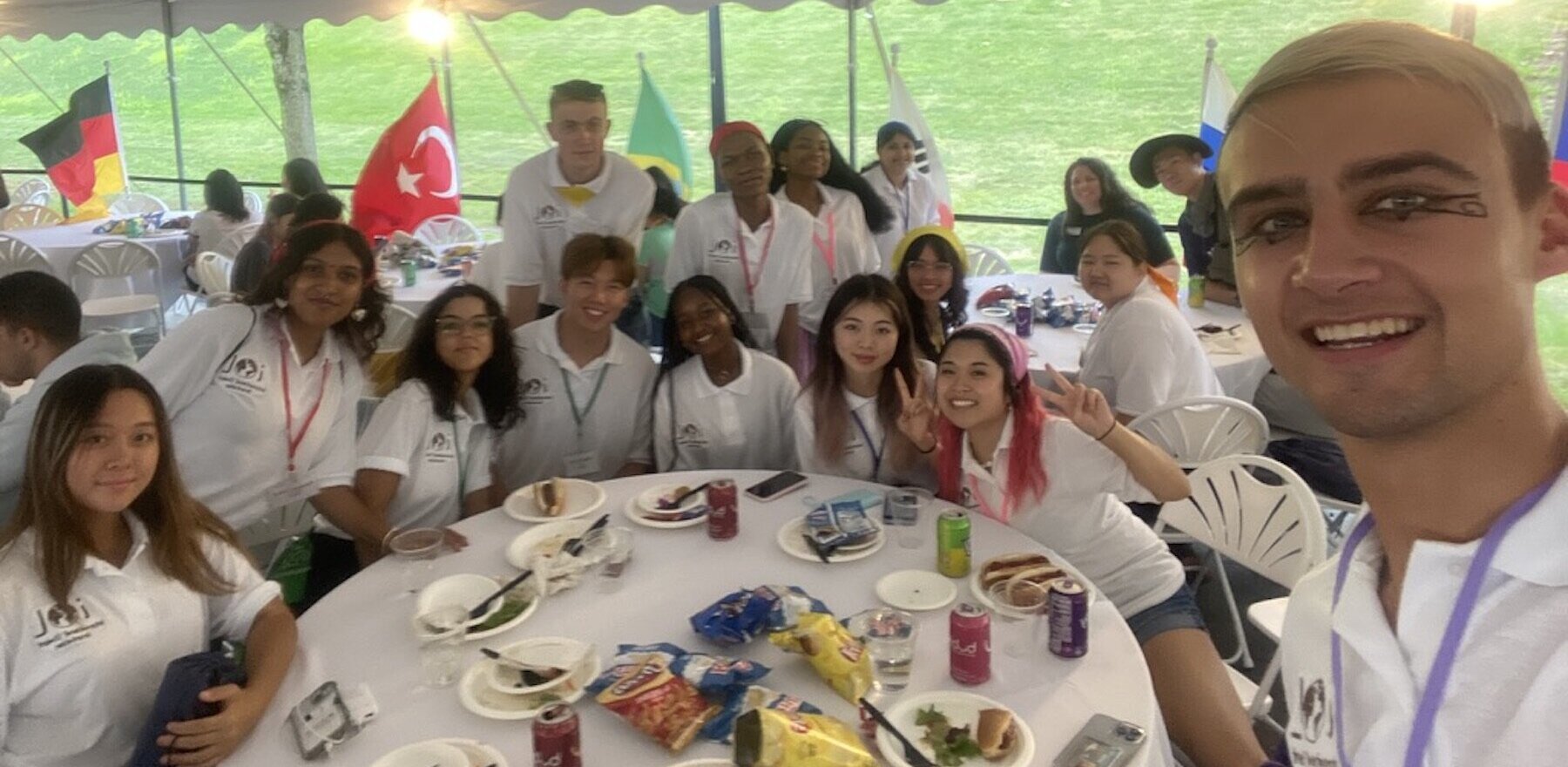 A person in the foreground taking a selfie of himself and several young people sitting at a table under an outdoor canopy