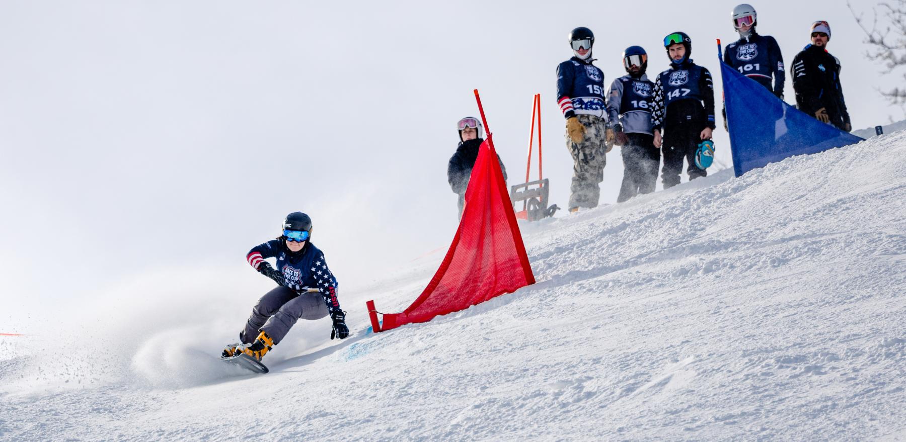 A photo of Mika Fizuka snowboarding down a snowy hill past a red flag while six people watch from above her