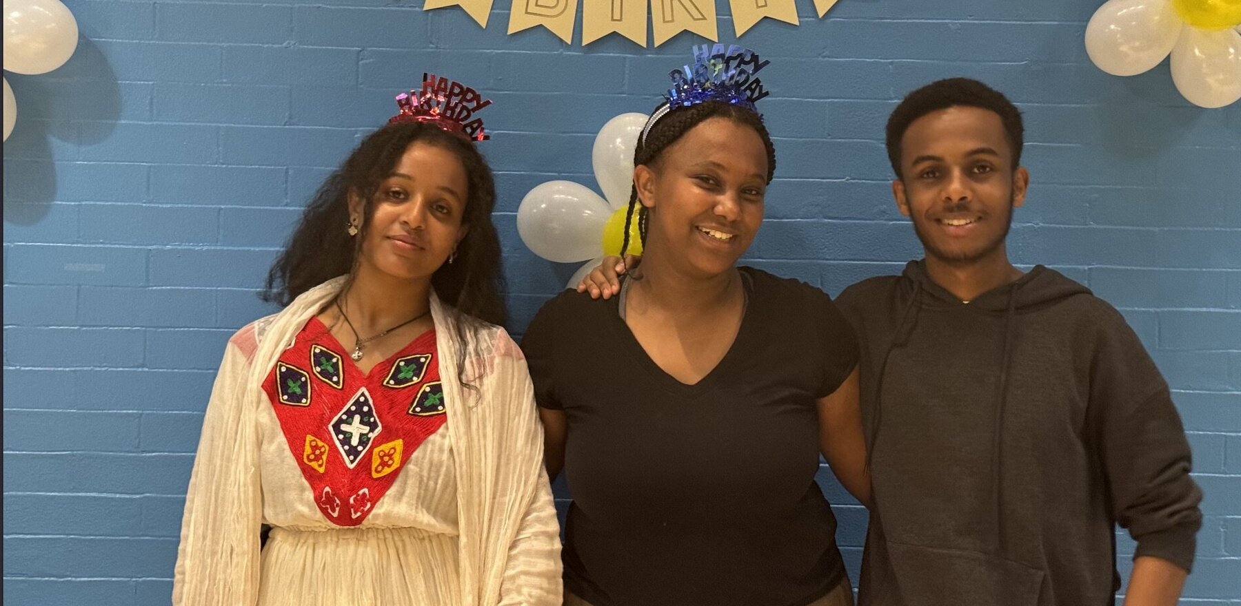 Three young people standing underneath a birthday banner and smiling for the camera