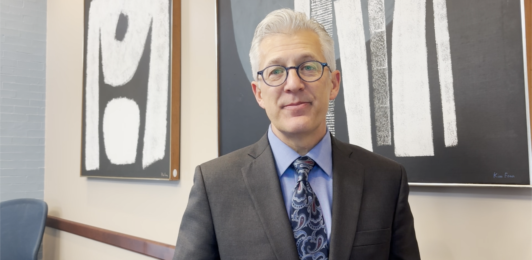 A man wearing glasses and a gray suit with a blue shirt and tie staring at the camera