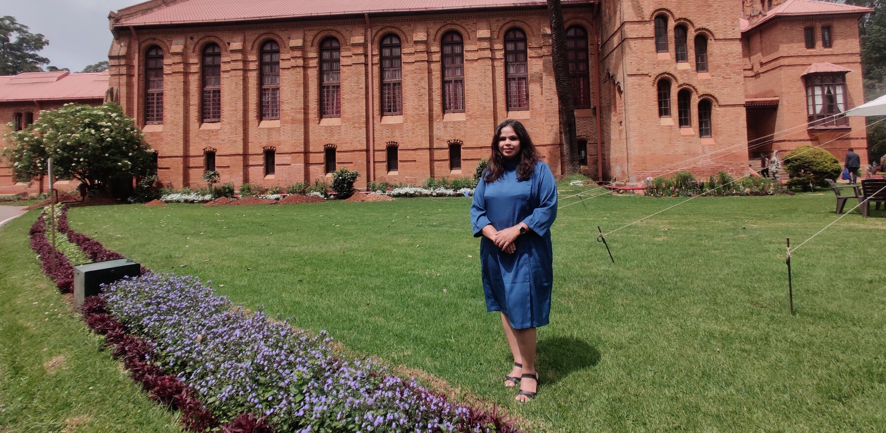 A woman in a blue dress stands with arms folded on the grassy front yard of a building