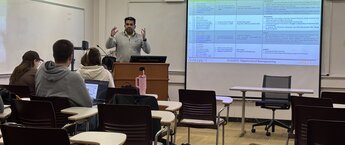 A teacher speaks to students from a podium in a classroom