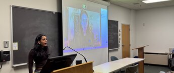 A young woman speaks at a podium while another woman speaks on an overhead projector screen behind her