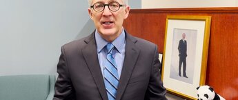 A man wearing glasses and a gray suit with a blue shirt and tie staring at the camera