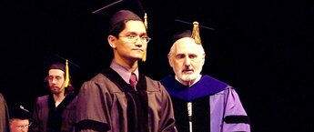 A young Thai man wearing a graduation robe, standing on a stage during a commencement ceremony