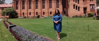 A woman in a blue dress stands with arms folded on the grassy front yard of a building