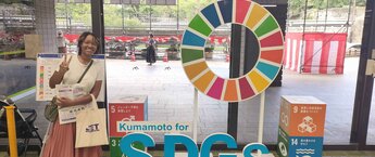 Zena Meighan standing in front of a display that says Kumamoto for SDGs