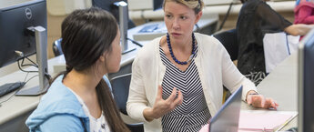 A woman speaking to another woman while sitting by some computers