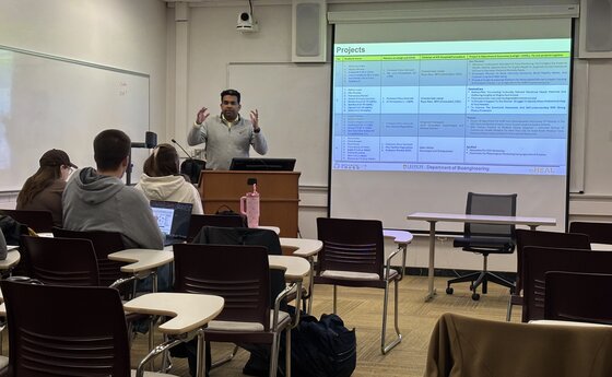 A teacher speaks to students from a podium in a classroom