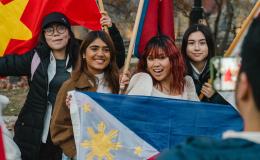 A man with his back to the camera takes a smartphone photo of four young owmen smiling and holding up a variety of flags