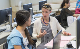 A woman talking to another woman while they sit in front of a table with computers