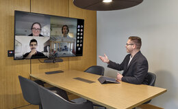 A man sitting at a table, talking to four people in a Zoom on a wall-mounted screen